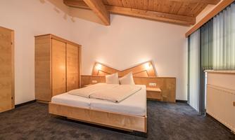 A modern bedroom with a wooden bed and compact furniture. The room has large windows and an elegant wooden ceiling.