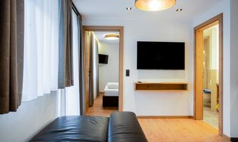 A modern room with a black sofa, a TV, and wooden flooring. In the background, additional rooms can be seen.