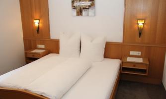 A cozy bedroom with a double bed and large pillows. The walls feature simple wooden shelves and plain wall lamps.