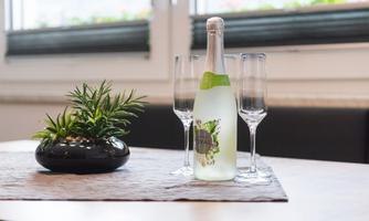 A bottle of sparkling drink stands on a table, surrounded by three champagne glasses. Next to the bottle is a small plant in a black pot.