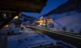 A picturesque winter landscape at night, with snow-covered mountains and illuminated houses. The road winds through the quiet, snowy surroundings.