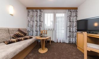 A cozy living space with a beige sofa, a small table, and a television. The window is decorated with white curtains and lets in a lot of light.