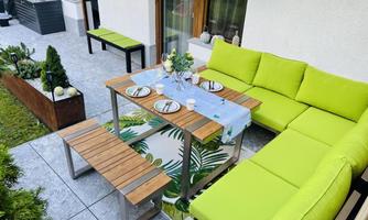 An inviting outdoor dining area with a green couch and a wooden table. The table is set and surrounded by plants on a paved floor.