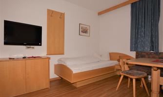A simple, modern room with a bed, table, and chairs. A television hangs on the wall, and there is a window with blue curtains.
