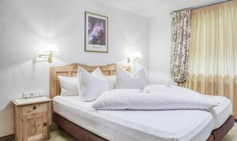 A cozy bedroom with a large double bed and two pillows. The walls are light and the lighting is soft; a window with curtains offers an inviting view outside.