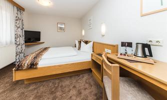 A cozy hotel room with a double bed and a desk. The room is bright and modernly furnished, with a television and a window for natural light.