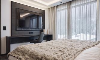 A modern bedroom with a large bed and a TV on the wall. Large windows let in a lot of light and offer a view of the snowy landscape.