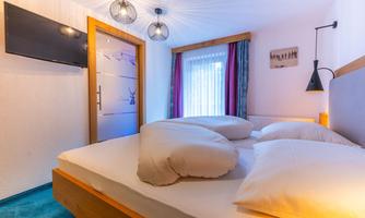 A cozy bedroom with a large bed and soft pillows. The walls are light, and there is a television as well as a window with curtains.