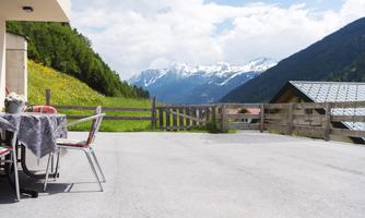 A terrace with garden furniture looks out over a stunning mountain landscape. In the background, snow-capped peaks and green meadows are visible.