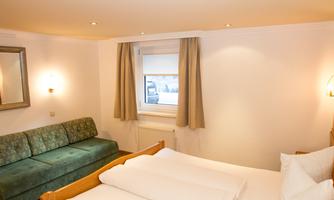 A cozy hotel room with a double bed and a green sofa. The room has large windows and warm light.