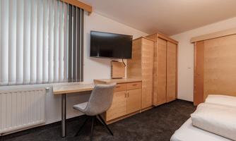 A modern room with a desk, a TV, and a wardrobe. The walls are light and equipped with vertical blinds.