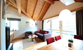 A modern living room with wooden beam ceiling and large windows. A cozy seating area and a dining table create an inviting atmosphere.