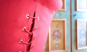 A red corset with eyelets and lacing. In the background, decorative pictures can be seen on the wall.