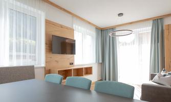 A modern living room with a large window and plenty of natural light. There is a TV, a dining table, and cozy furniture in bright colors.