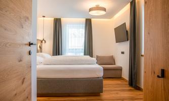 A modern bedroom with a double bed and a cozy sofa. Large windows let in plenty of light and the walls are painted in neutral colors.