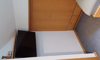 A simple room with a television on the wall and a wooden wardrobe. The floor is carpeted and there is a bed with gray bedding.