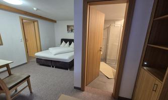 A simple room with a double bed and a table. In the background, a bathroom with a glass shower is visible.