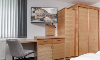 A modern room with a desk, a chair, and a television on the wall. In the background, there is a wooden wardrobe.
