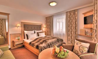 A cozy hotel room with a double bed and comfortable furniture. There is a table with fresh fruit and large windows that let in a lot of light.