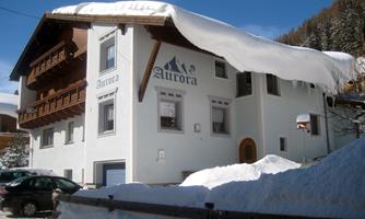 A cozy hotel named "Aurora" with a snow-covered roof. The façade is white and well-preserved, surrounded by snow.