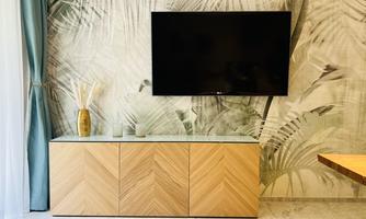 A modern living space with a flat-screen TV on the wall. In front of the TV is a light wooden commode, and in the background are plant motifs.