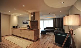 A modern kitchen-living area with wooden surfaces and elegant lighting. The living area is cozy with a sofa and a large window.