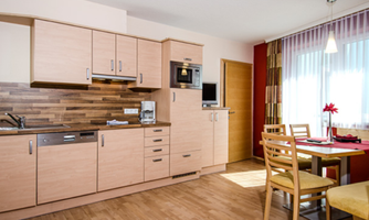 A modern kitchen with light wood cabinets and stainless steel appliances. There is a small dining area with a table and chairs.