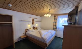 A cozy bedroom with a double bed and wooden ceiling. The room is bright and friendly, with a window and a few furniture pieces.