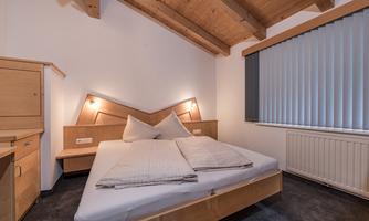 A modern bedroom with a wooden ceiling and a large bed. The walls are simple, and there are windows with blinds.
