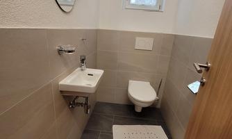A modern bathroom with a white wall, a sink, and a toilet. The floor is tiled in gray, and a large mirror is mounted on the wall.