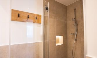 A modern shower with glass partition and high-quality tiles. On the wall, there is a wooden shelf with hooks.