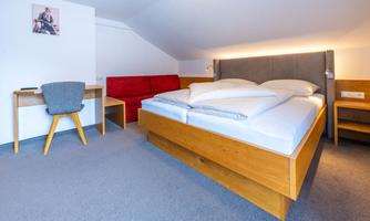 A cozy bedroom with a large bed, a desk, and a red sofa. The room is bright and modernly furnished.