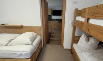 A bright room with a single bed and a wooden bunk bed. The door leads to a small kitchen in the background.