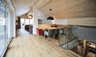 A modern, light-flooded living room with wooden walls and a large table. The room has large windows and an open atmosphere.
