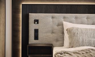 A modern bedroom with an upholstered headboard and soft lighting. Next to the bed is a nightstand with controls.