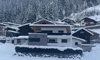 A modern house in the snow, surrounded by snow-covered mountains and pine trees. The landscape is wintry and idyllic.