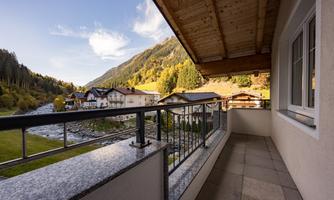 A balcony with a view of a picturesque landscape. In the background, mountains, trees, and several houses can be seen.