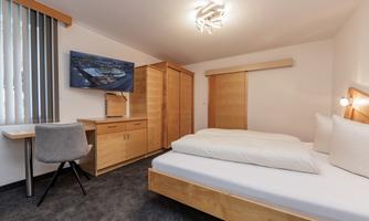 A modern room with a large bed and wooden furniture. There is a desk, a chair, and a TV on the wall.