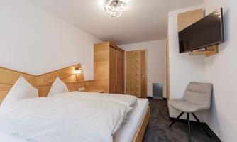 A modern room with a double bed and wooden furniture. There is a television, a chair, and plenty of daylight.