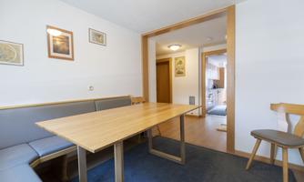 A simple dining area with a wood-colored table and comfortable benches. The walls are decorated with pictures, and a door leads to the adjacent kitchen.