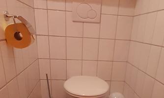 A small, simple bathroom with white tiles and a toilet. Hanging on the wall are toilet paper and a trash can.