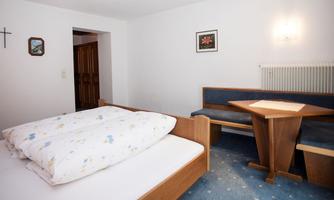 A simple, bright bedroom with a double bed and a small dining table. The walls are kept neutral, and there are some decorative elements.