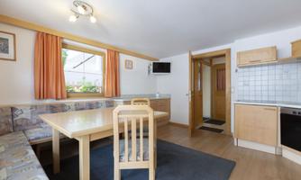 A cozy dining area with a wooden table and chairs. The kitchen is bright and well-equipped, with a window and orange curtains.