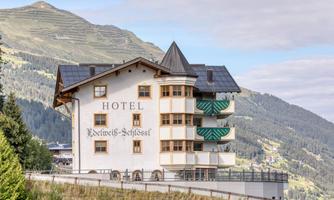 A picturesque hotel in the mountains with several balconies and a typical alpine style. In the background, green meadows and mountains are visible.