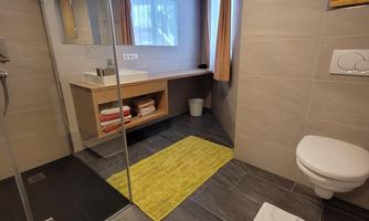 A modern bathroom with a shower, a sink, and a toilet. The floor is decorated with gray tiles and a yellow rug.
