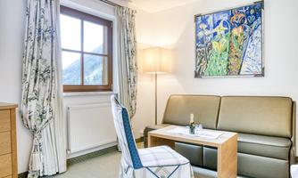 A cozy living area with a sofa and a table. A colorful painting hangs on the wall and a window lets in plenty of daylight.