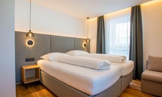A modern bedroom with a large double bed and a cozy seating area. The walls are bright and there is soft lighting.