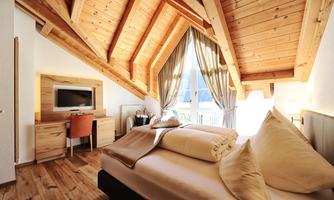 A cozy bedroom with a wooden ceiling and a large window. The room is bright and well-furnished with a bed and a desk.