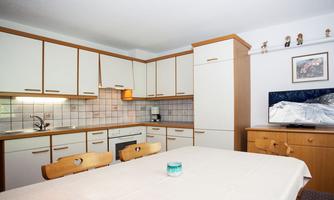 A modern kitchen with wooden cabinets and a large workspace. The dining table is simply set, and a TV is in the background.