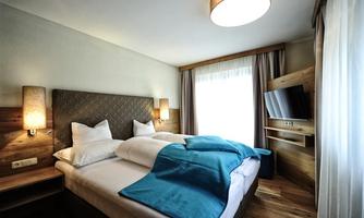 A modern hotel room with a double bed and wooden details. The windows let in plenty of daylight, and there is a television on the wall.